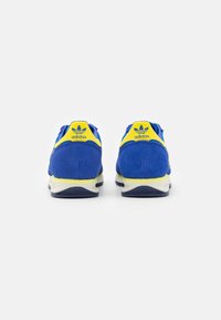 Baskets en suède bleu avec des accents jaunes et une semelle blanche. Présente le logo Adidas au niveau du talon et de la languette, conçues pour une utilisation décontractée.