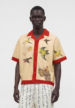 Personne à la peau foncée portant une chemise beige avec une bordure rouge et des oiseaux colorés portant un panier, associée à un pantalon blanc avec du texte manuscrit bleu.