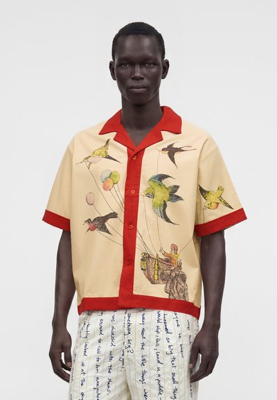 Personne à la peau foncée portant une chemise beige avec une bordure rouge et des oiseaux colorés portant un panier, associée à un pantalon blanc avec du texte manuscrit bleu.