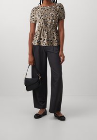 Donna che indossa una maglietta beige con stampa leopardata a maniche corte, pantaloni neri a gamba larga, ballerine nere, mentre tiene una borsa nera con dettagli dorati.
