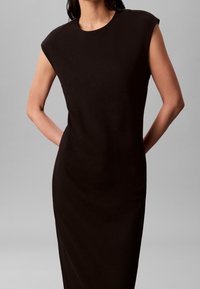 Femme portant une robe noire cintrée sans manches avec de larges épaules, debout les mains derrière le dos sur un fond uni.