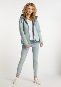 Ljusblå, texturerad fleecejacka med huva, dragkedja fram, snörningsdetaljer, bärs över en vit skjorta och ljusa jeans.