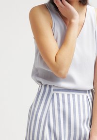 Světle šedý bezrukávový top z jemné látky, který je spárován s bílými a modrými pruhovanými kalhotami s vysokým pasem, majícími dobře padnoucí střih a hladkou texturu.