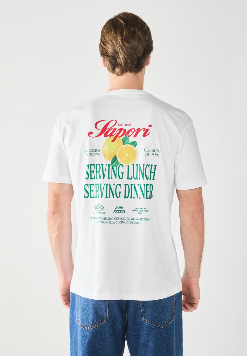 Hvid bomulds T-shirt med farverig tekst og grafik, herunder citroner og detaljer om en restaurants frokost- og middagsservice.