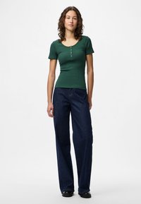 Top curto de mangas curtas em verde escuro com decote arredondado e detalhe de botão, combinado com calças de ganga soltas em azul escuro e sapatos pretos.