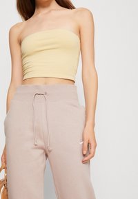 Kvinna iförd en beige bandeau crop top och ljus mauve joggbyxor med dragsko, som håller en liten tan väska mot en enkel bakgrund.