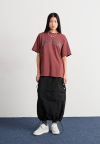 Camiseta oversize de color burdeos de Nike con logo gris, combinada con una falda larga negra con bolsillos cargo y tobillos ajustados. Zapatillas blancas.