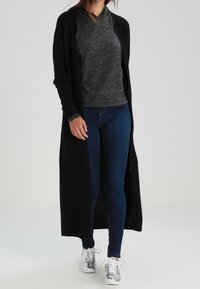 Długi czarny kardigan na szarym swetrze, w połączeniu z ciemnoniebieskimi dżinsami i srebrnymi tenisówkami. Kardigan ma luźny krój i otwarty przód.