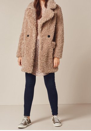 Tøff beige frakke med dobbeltradet design, store revers og forlommer, båret over en mønstret kjole og mørke jeans med sneakers.