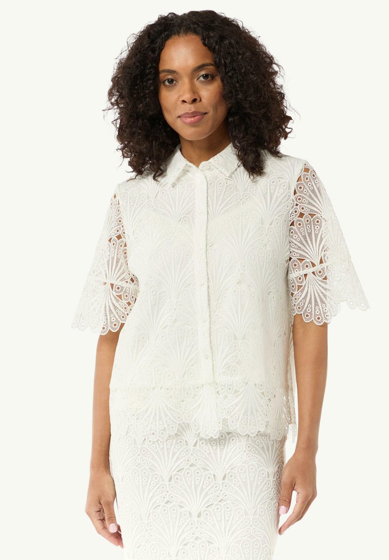 Weiße Spitzenbluse mit kurzen, gewellten Ärmeln. Verfügt über ein florales Muster, Knopfverschluss und strukturierte Stoffqualität. Das mehrlagige Design erzeugt Tiefe.