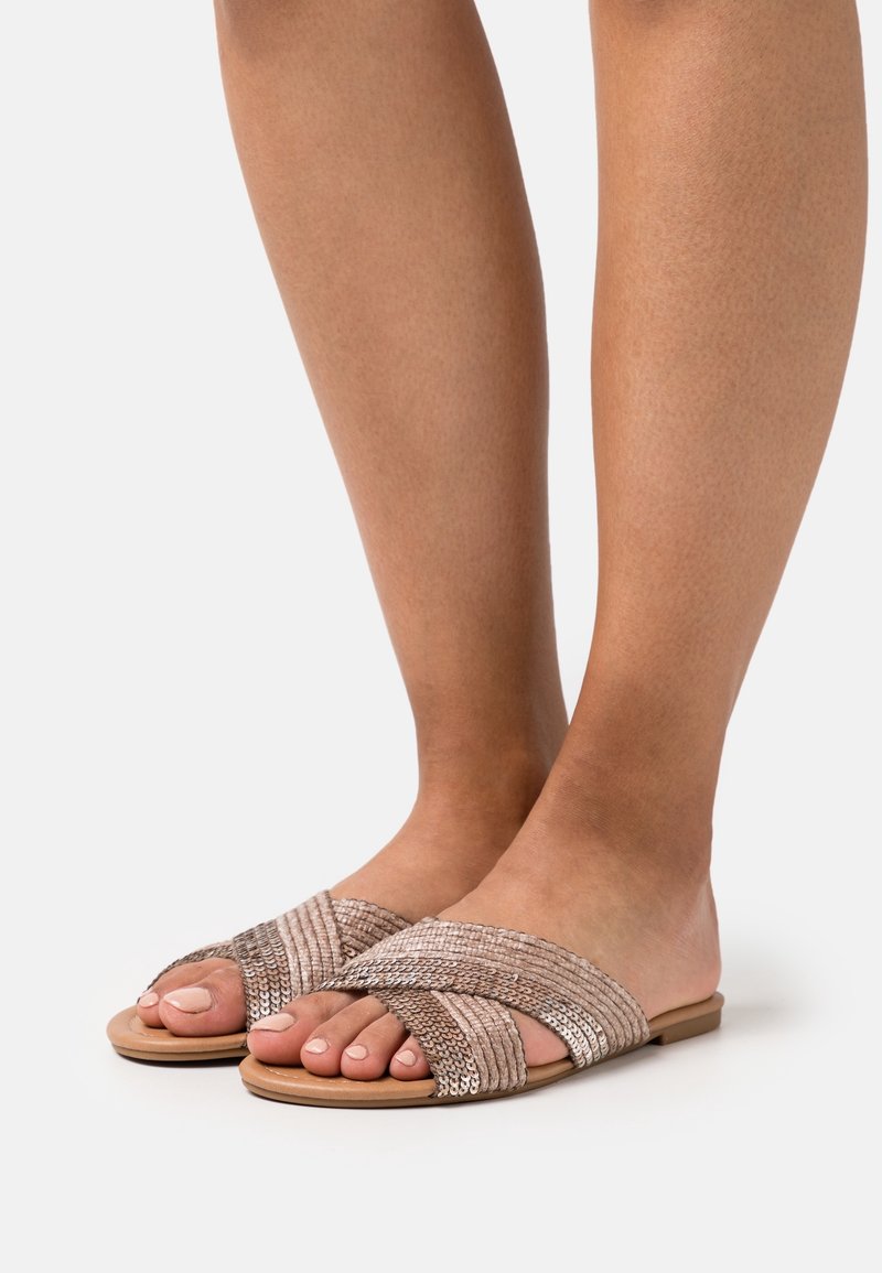 Sandales à glissière beiges avec des brides croisées tissées, présentant une finition texturée et des semelles plates. Design à bout ouvert révélant des ongles de pieds manucurés.