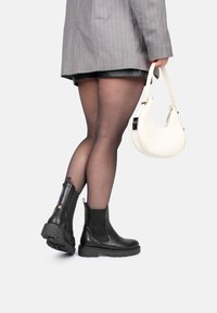 Botines negros de cuero con paneles elásticos en los laterales, suelas de goma gruesas y una lengüeta roja. Llevados con medias negras transparentes y un blazer gris.