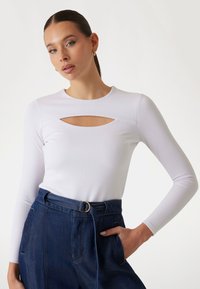 White ribbed long-sleeve top with a front cutout. Paired with high-waisted blue denim pants, featuring a belt with a gold buckle.