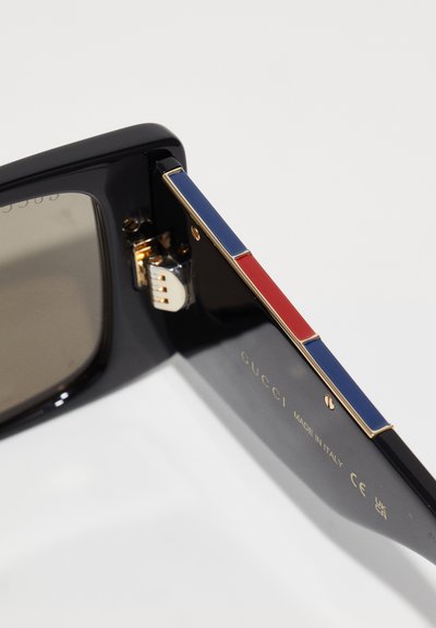 Črne sončne očala z gunmetal opremo, ki imajo modre in rdeče akcentne barve na nosilcih. "GUCCI" vgravirano v zlati barvi blizu tečaja.