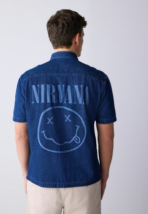 Mand iført kortærmet, blå denimskjorte med "NIRVANA" og et smilende ansigt printet på ryggen, lyse bukser, vendt væk fra kameraet.