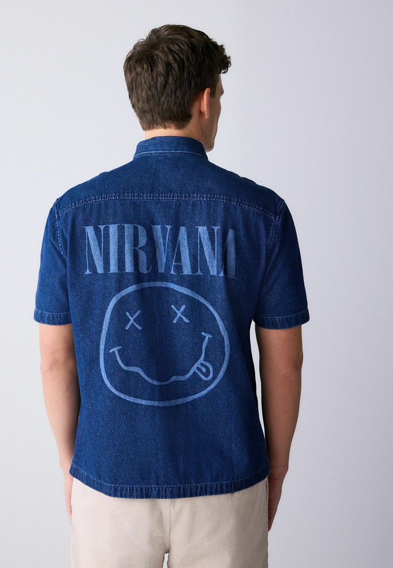 Homme portant une chemise en denim bleu à manches courtes avec l'inscription « NIRVANA » et un visage souriant graphique au dos, pantalon de couleur claire, de dos.