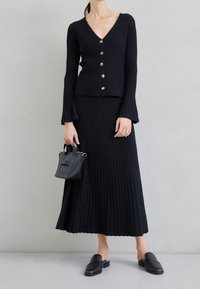 Černý žebrovaný pletený cardigan s výstřihem do V a zlatými knoflíky, doplněný o dlouhou sukni ve stejném stylu. Černé nazouvací boty a malá kabelka dokončují vzhled.