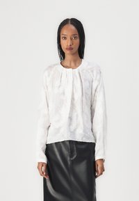 Hvit brodert bluse med lange ermer, rund halslinning og folder, kombinert med et svart skinnskjørt. Glatt teksturert stoff.