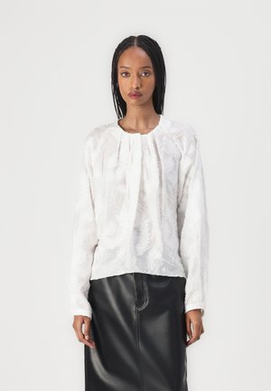 Hvit brodert bluse med lange ermer, rund halslinning og folder, kombinert med et svart skinnskjørt. Glatt teksturert stoff.