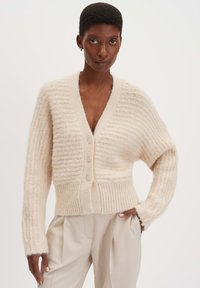 Beige gestrickte Strickjacke mit geripptem Textur, V-Ausschnitt und drei vorderen Knöpfen, mit langen Ärmeln und einer taillierten Saum. Glattes, weiches Material.