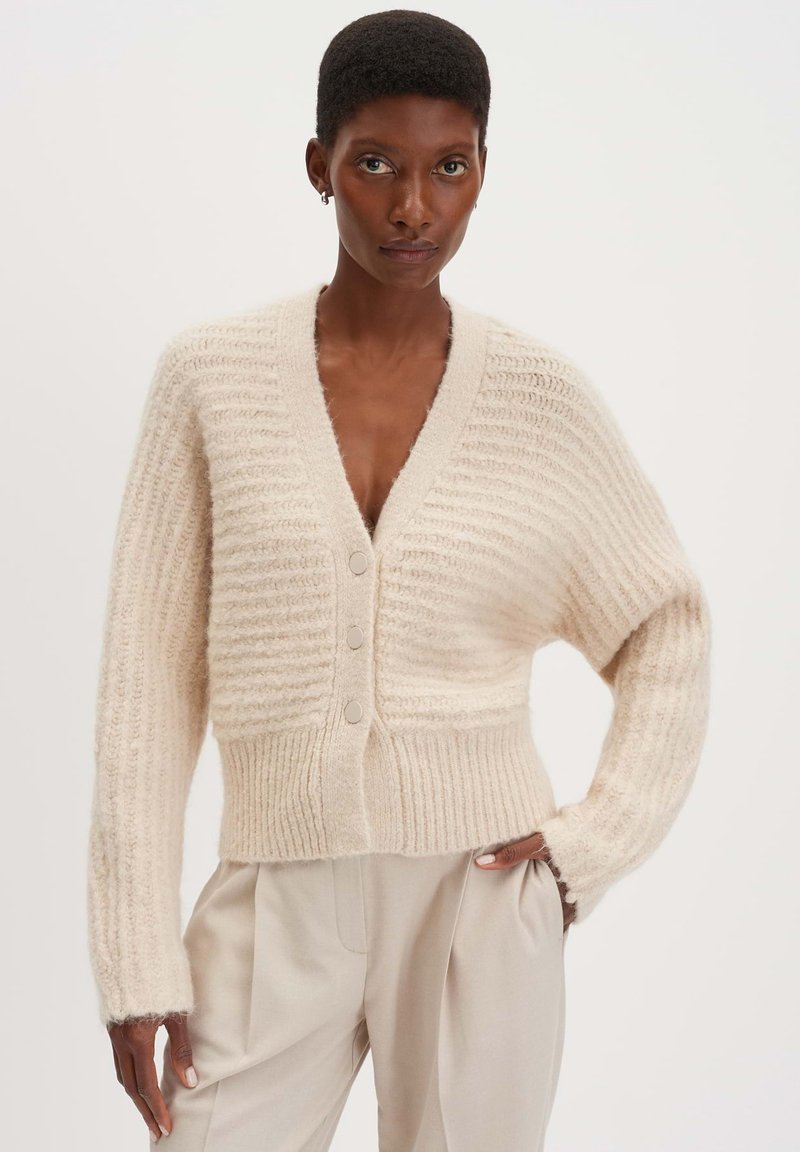 Beige gestrickte Strickjacke mit geripptem Textur, V-Ausschnitt und drei vorderen Knöpfen, mit langen Ärmeln und einer taillierten Saum. Glattes, weiches Material.