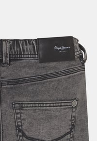 Szare dżinsy z tyln�ą kieszenią, czarna skórzana metka z logo "Pepe Jeans London" oraz widoczną pętelką na pasek na białym tle.