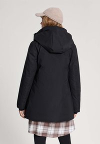 Kobieta w czarnym płaszczem z kapturem, beżowej dzianinowej czapce i brązowej spódnicy w kratę, widoczna od tyłu na jednolitym jasnym tle.