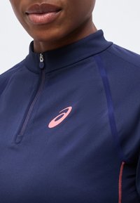 Κοντινό πλάνο ενός ατόμου που φορά ναυτικό μπλε αθλητικό τοπ με φερμουάρ μέχρι τη μέση, με ροζ λογότυπο Asics στο στήθος και διακριτική ροζ επένδυση στο πλάι.