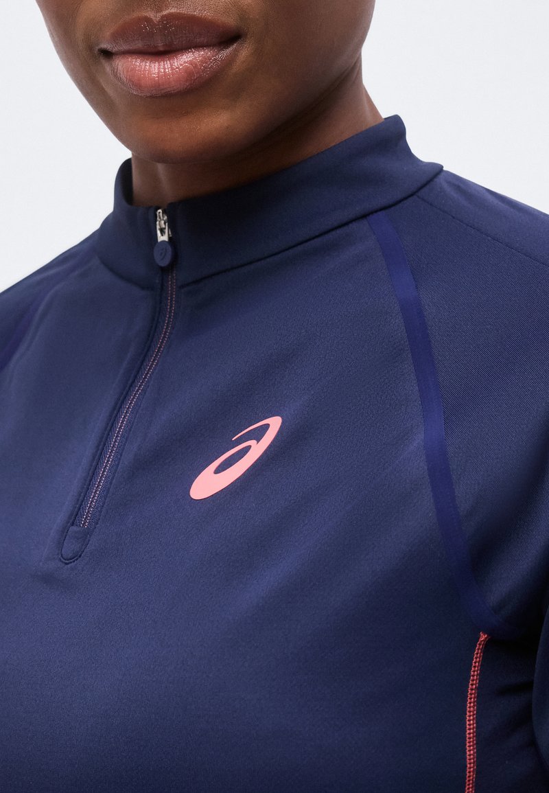 Close-up af person iført navyblå atletisk quarter-zip top med lyserødt Asics-logo på brystet og diskrete lyserøde syninger i siden.