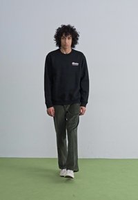 Felpa nera con logo, pantaloni in velluto a coste verde oliva e sneaker bianche su un pavimento verde contro uno sfondo minimalista.