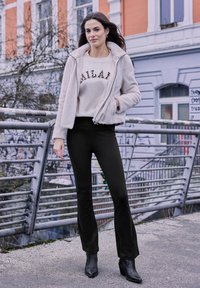 Veste en fausse fourrure beige clair, top crème à manches longues avec impression "MILAN", pantalon noir et bottines, posés sur un pont.