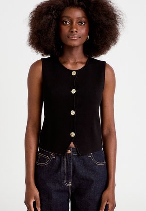 Vrouw met volumineus krullend haar, gekleed in een mouwloze zwarte top met knopen en donkere spijkerbroek, tegen een effen achtergrond.