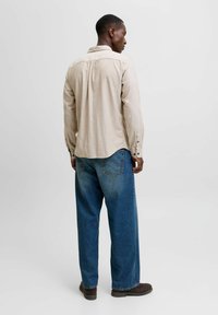 Man wearing beige long-sleeve shirt, loose blue jeans, and dark shoes, standing facing away against a plain light background.