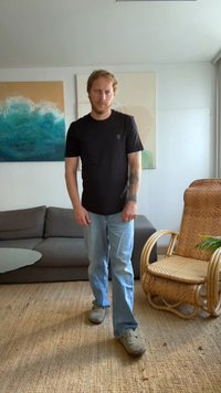 Homem com tatuagens a usar uma camisa preta e calças de ganga claras a andar sobre um tapete tecido na sala de estar, com um sofá castanho e uma cadeira de vime.