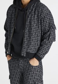 Giacca nera con cerniera e motivo a testo ripetuto, abbinata a una felpa nera con cappuccio. Presenta polsini elasticizzati e una vestibilità comoda, realizzata in materiale strutturato.