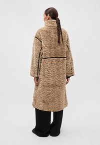 Garšs, teksturēts bēšs mētelis ar pūkainu apdari, melniem akcentiem un zemu apkakli. Mētelis ir brīva piegriezuma un sasniedz vidus teļa līmeni.