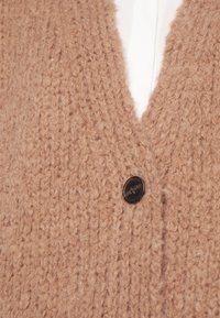 Beige gestrickte Strickjacke mit V-Ausschnitt und strukturiertem Gewebe, mit einem einzelnen schwarzen Knopfdetail in der Nähe des Ausschnitts.