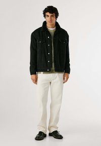 Hombre vestido con chaqueta negra de cuello de borreguillo, suéter de punto verde oliva, pantalones crema y zapatos de cuero negros, de pie frente a un fondo blanco.