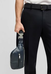 Borsa in pelle blu marino con finitura lucida, chiusura con zip e tracolla strutturata tenuta in una mano. Abbinata a pantaloni neri su misura.