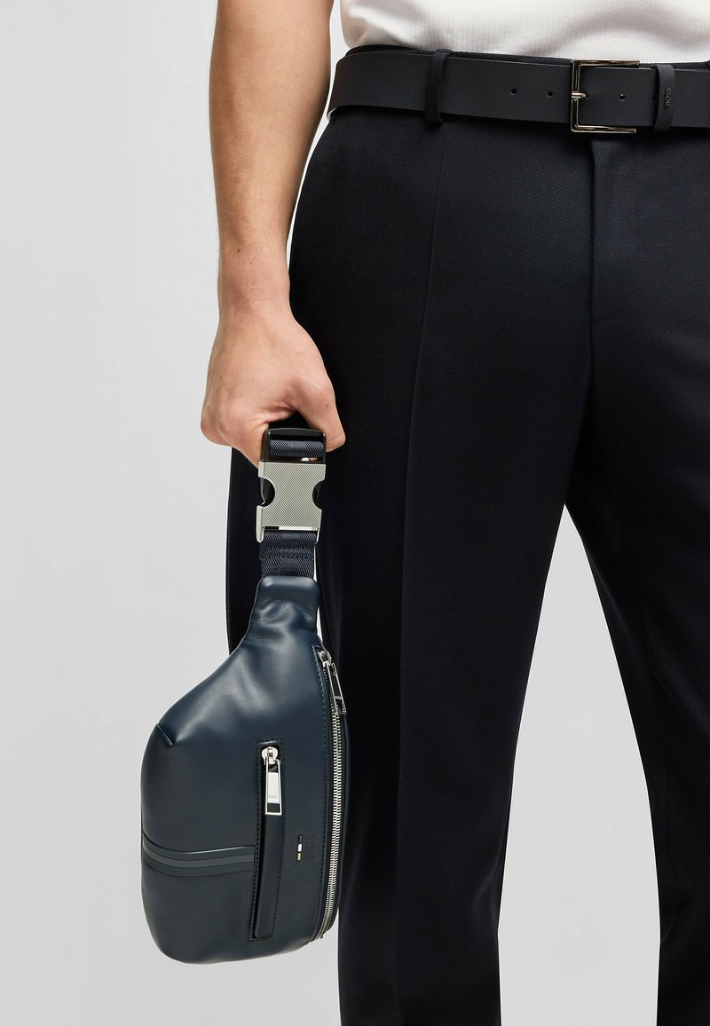 Borsa in pelle blu marino con finitura lucida, chiusura con zip e tracolla strutturata tenuta in una mano. Abbinata a pantaloni neri su misura.