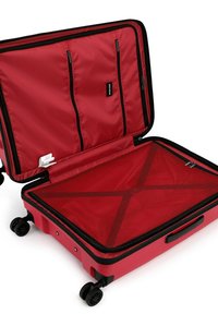Červený tvrdý kufr s černými akcenty, do kterého se vstupuje shora, má dvě kapsy na zip a křížový popruh pro zajištění obsahu.