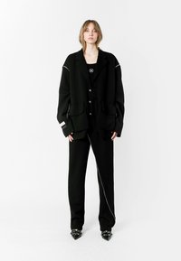Blazer negro de corte oversize con dos bolsillos delanteros, combinado con pantalones negros ajustados que presentan un detalle de cremallera diagonal. Llevado con tacones de punta.