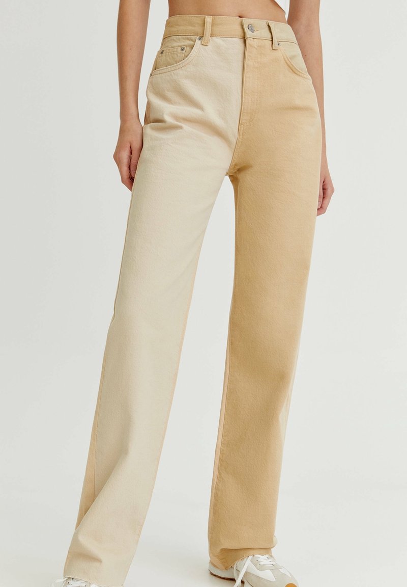 Femme portant un jean droit taille haute avec une jambe beige et l'autre couleur sable clair, associé à des baskets blanches.