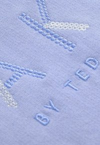 Světle modrá látka s texturovanou výšivkou a hladkým povrchem, zobrazující název značky "by Ted" a dekorativní vzor.