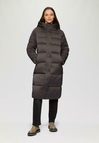 Doudoune longue marron foncé avec capuche, présentant un design matelassé, des boutons sur le devant et des poches latérales. Portée sur un pantalon noir et des bottes.