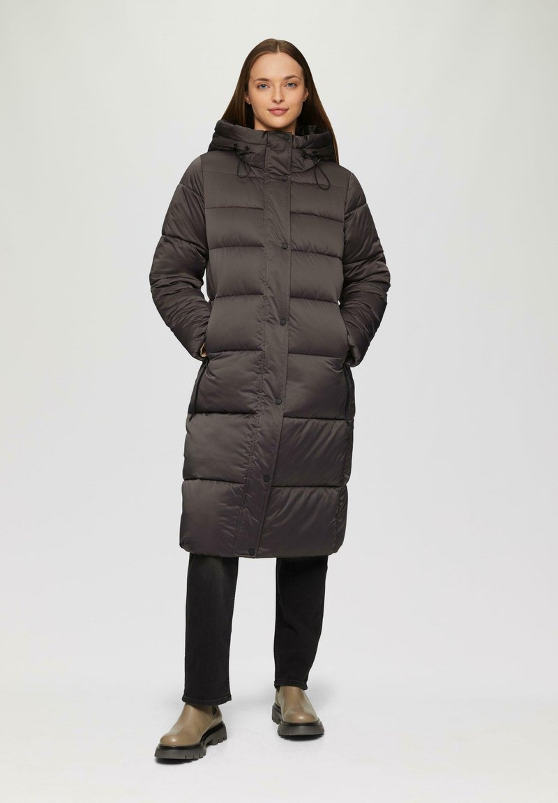 Doudoune longue marron foncé avec capuche, présentant un design matelassé, des boutons sur le devant et des poches latérales. Portée sur un pantalon noir et des bottes.