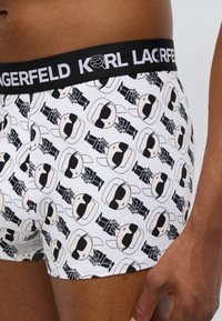 Baltos bokso šortai su juodu ir rudos spalvų kartotiniu animaciniu atspaudu, vaizduojančiu figūrą su akiniais nuo saulės, turintį juodą juosmenį su užrašu „KARL LAGERFELD“.