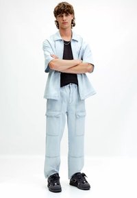 Svetlomodrá krátka košeľa na gombíky cez čierny top, skombinovaná so svetlomodrými cargo nohavicami a čiernymi teniskami. V stoji, s prekríženými rukami.