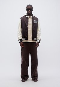 Άνδρας που φοράει μαύρο και λευκό varsity μπουφάν με το λογότυπο NY, μπεζ πουλόβερ, φαρδιά μαύρη παντελόνια, σκούρα παπούτσια και μαύρα γυαλιά ηλίου, στέκεται μπροστά σε λευκό φόντο.