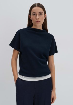 Navy blue textured top with short sleeves and a high neckline, layered over a white hem, paired with dark trousers.